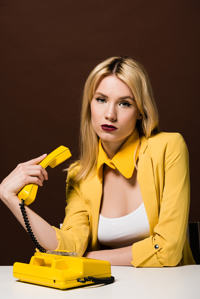hermosa chica rubia elegante sosteniendo el auricular amarillo y mirando a la cámara en marrón
 - Foto, Imagen