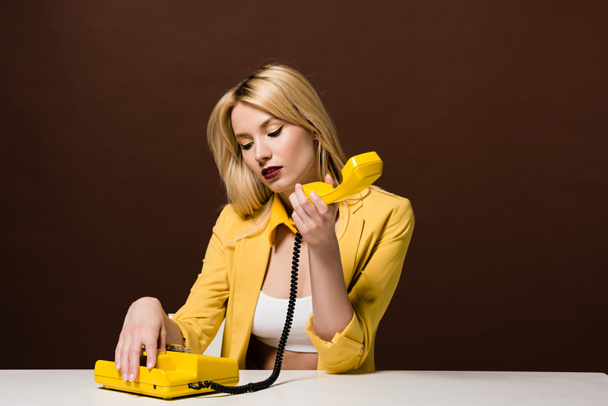 pensive blonde girl holding yellow vintage telephone while sitting on brown  - Фото, зображення