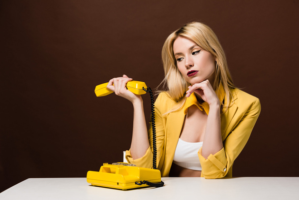 nachdenkliches blondes Mädchen mit gelbem Hörer, während es auf braun sitzt  - Foto, Bild