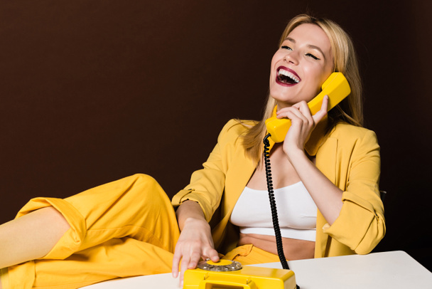 fröhliches blondes Mädchen, das mit gelbem Vintage-Telefon spricht und über braun lacht - Foto, Bild