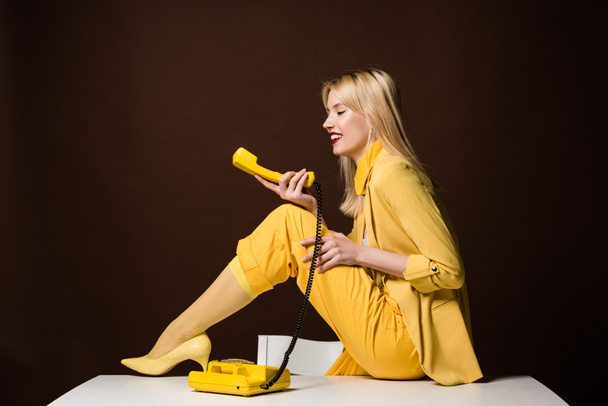 side view of beautiful stylish blonde girl holding yellow vintage phone and smiling on brown - Foto, Bild