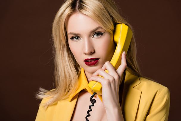 atractiva mujer rubia hablando por teléfono vintage amarillo y mirando a la cámara aislada en marrón
  - Foto, Imagen