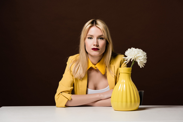 kaunis tyylikäs blondi tyttö katselee kameraa istuessaan pöydässä valkoinen kukka keltainen maljakko ruskea
 - Valokuva, kuva