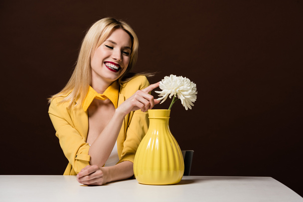 beautiful smiling blonde woman in stylish yellow outfit touching white flower in vase on brown  - Фото, изображение