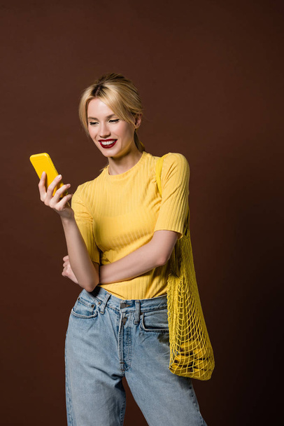 beautiful smiling blonde girl with string bag using yellow smartphone isolated on brown - Fotoğraf, Görsel