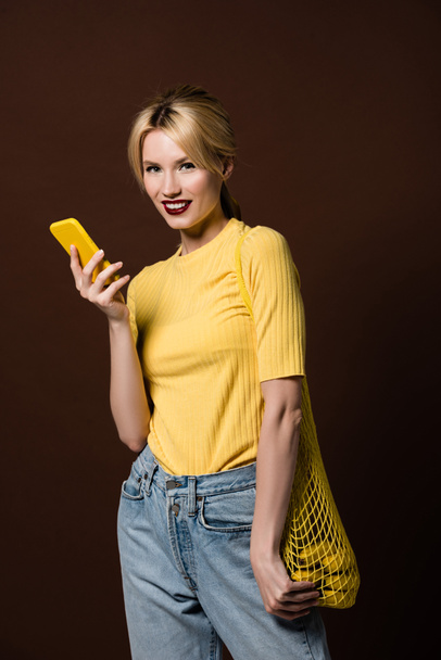 beautiful stylish blonde girl with bananas in string bag using yellow smartphone and smiling at camera isolated on brown - Foto, Imagen