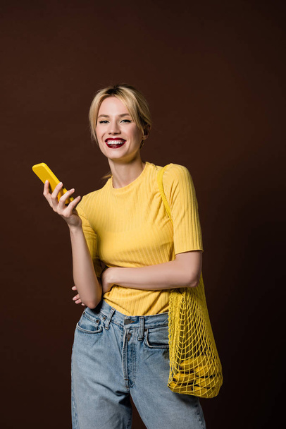 cheerful stylish blonde girl with bananas in string bag using yellow smartphone and smiling at camera isolated on brown - Valokuva, kuva