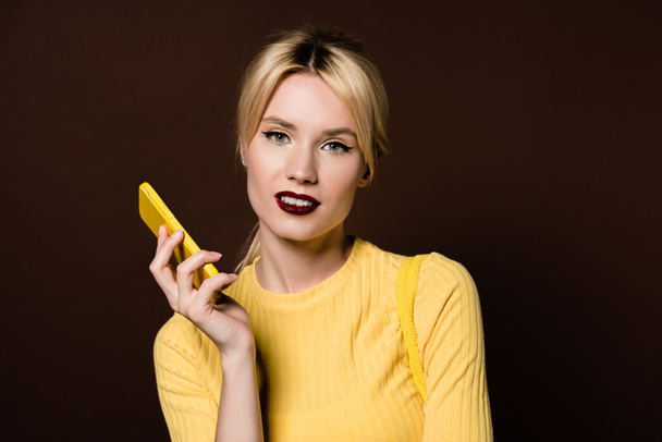 beautiful stylish blonde girl holding yellow smartphone and smiling at camera isolated on brown - Fotoğraf, Görsel