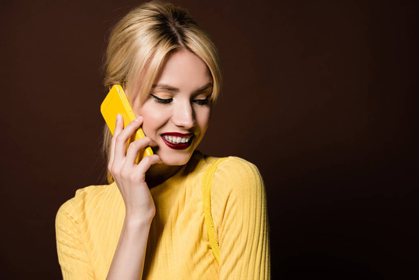 schöne lächelnde blonde Mädchen im Gespräch mit gelben Smartphone isoliert auf braun - Foto, Bild
