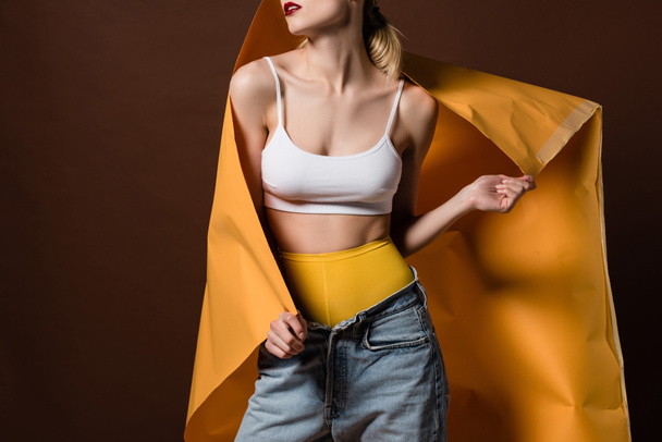 cropped shot of seductive stylish girl posing with orange paper isolated on brown - Foto, afbeelding