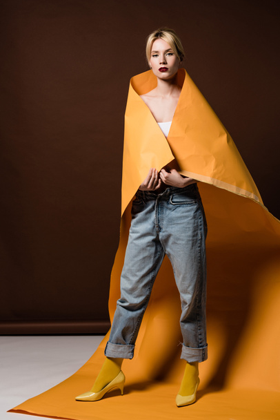 full length view of beautiful stylish blonde girl wrapped in orange paper looking at camera on brown  - Foto, immagini