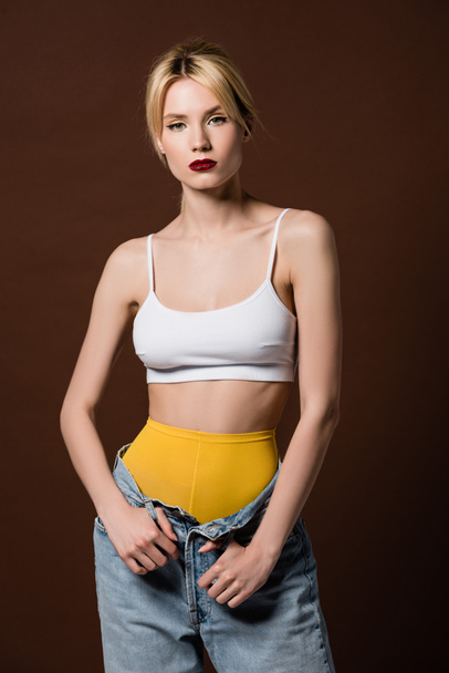 portrait of beautiful blonde woman in yellow tights and jeans looking at camera isolated on brown  - Foto, Bild