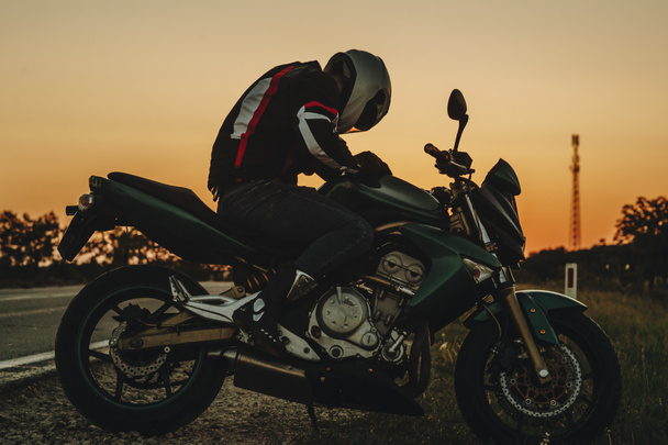 Вид сбоку на удивительный кавказский мотоциклист, путешествующий на своем велосипеде во время отдыха, сидя перед закатом на дороге
. - Фото, изображение