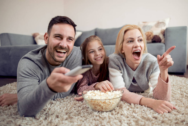 Father mother and daughter lying on floor eating popcorn and watching TV - Фото, изображение