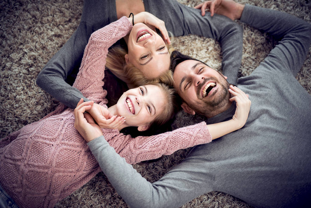 Вид сверху на молодую семью, которая смотрит в камеру и улыбается, лежа на полу дома
 - Фото, изображение
