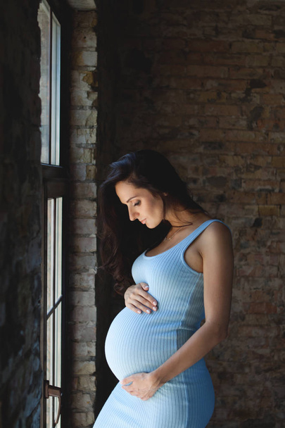 Kobiety w ciąży brunetka w niebieskiej sukience przytulił jej brzuch w ciąży. W ciąży kobieta stoi przy oknie - Zdjęcie, obraz