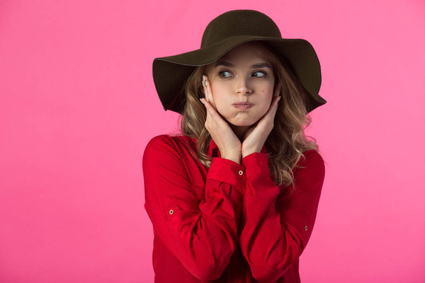 beautiful young girl in a red shirt and hat on a pink background with puffed cheeks - Photo, image
