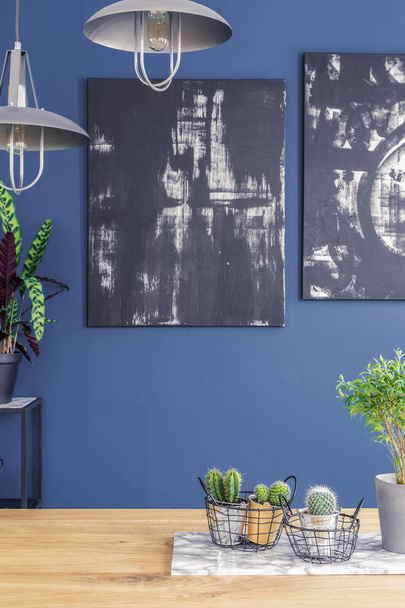 Black paintings on a blue wall and industrial pendant lights over a wooden kitchen interior table with plants and a marble board - Fotografie, Obrázek