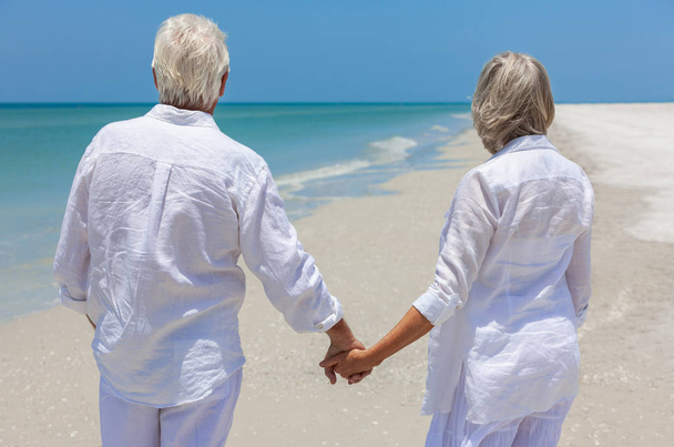 Вид ззаду щасливого старшого чоловіка і жінки пара ходить і тримає руки на пустельному тропічному пляжі з яскравим ясним блакитним небом
 - Фото, зображення