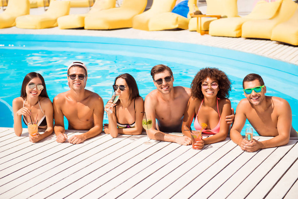 Six international hot teens students in diverse swim wear and spectacles are chatting in the pool, sunshine, blue sky, drinks are on white wooden floor, holiday together on beach resort - Фото, зображення