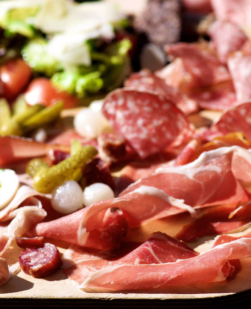 Wooden Plate of Delicious Cold Cuts with Smoked Ham, Cured Sausage, Salami, Marinated Onions and Vegetables closeup Outdoors. Focus on Foreground - Photo, Image