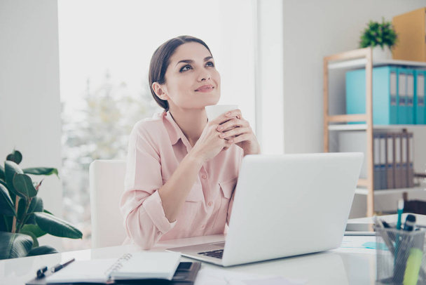 Dreams come true! Minded ponder charming executive secretary sitting in modern office holding mug with tea in hands looking up dreaming about vacation planning holiday having laptop on the table - Photo, image