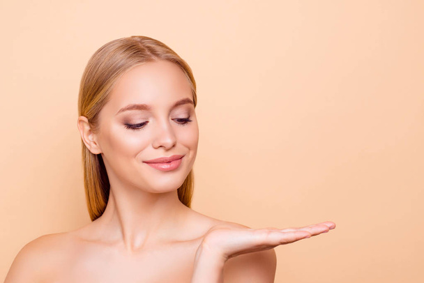 Cute charming nude natural pure girl with smooth soft skin looking at copy space empty place on her palm isolated on beige background hydratuin, moisturizing, nutrition advertisement concept - Photo, image