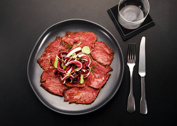 Sliced cold roast beef with fresh radicchio salad in a modern black table setting with utensils and wineglass viewed top down - Photo, Image