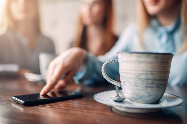Cup of coffee and mobile phone on wooden table, closeup. Girls leisures in cafe. Female friends sitting in restaurant on couch and drinks coffee, gossipers meeting - Fotó, kép