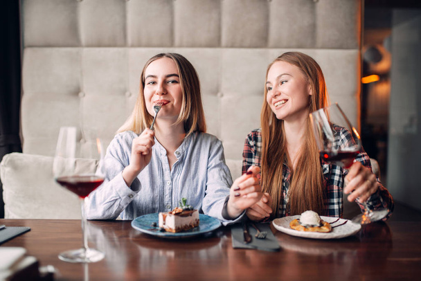 δύο χαμογελώντας νεαρές γυναίκες τρώνε γλυκά κέικ στο καφέ και πίνοντας κόκκινο κρασί  - Φωτογραφία, εικόνα