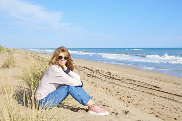Full length shot of middle aged woman sitting at the beach and relaxing.  - 写真・画像