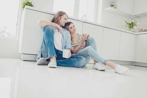 Low angle view of lovely romantic couple holding cups with coffee in hands enjoying weekend holiday together sitting on floor in modern white kitchen. Idyllic daydream harmony concept - Fotografie, Obrázek