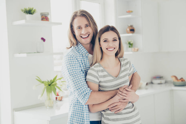 He vs she together forever! Portrait of positive toothy partners embracing in modern white kitchen with interior looking at camera. Daydream delight mood concept - Фото, изображение