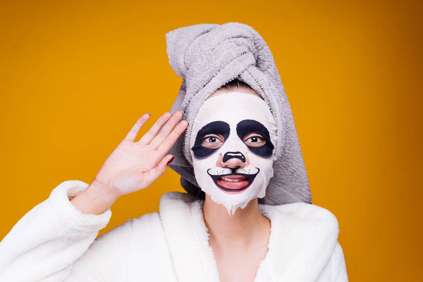 a nice young girl in a bathrobe and with a towel on her head takes care of herself, on her face mask with a panda face - Valokuva, kuva