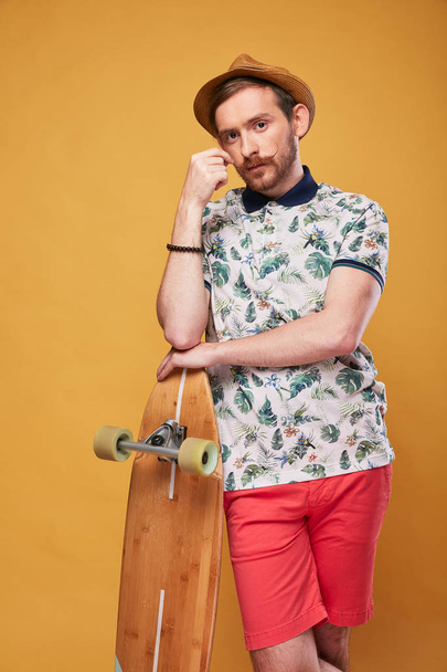 charismatic young man with handlebar mustache dressed in tropical print polo shirt, red shorts and straw hat, pointing on longboard in his hand, with longboard in another hand. Studio shot with yellow background  - Foto, Imagem