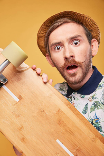 Astonished young man with handlebar mustache dressed in tropical print polo shirt and straw hat, with longboard in his hands. Studio shot with yellow background  - 写真・画像