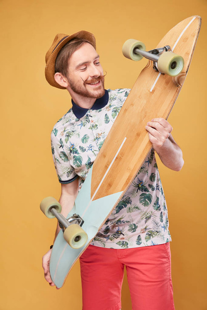Cheerful young man with handlebar mustache dressed in tropical print polo shirt, red shorts and straw hat, looks at the longboard in his hands. Studio shot with yellow background  - Fotoğraf, Görsel