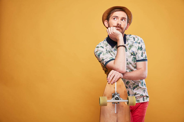 Pensive young man with handlebar mustache dressed in tropical print polo shirt, red shorts and straw hat, leaned on the longboard. Studio shot with yellow background  - Foto, afbeelding