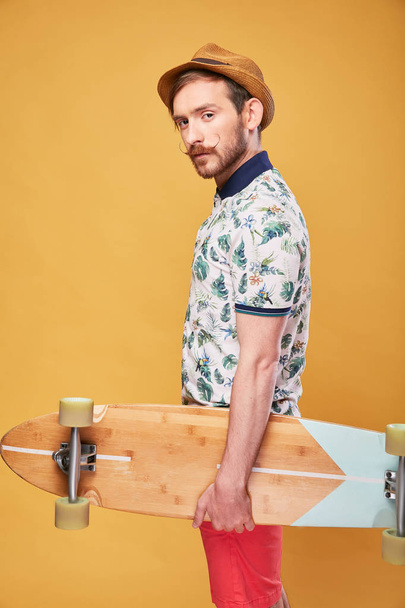 Bright portrait of young man with mustache and beard dressed in straw hat, tropical polo shirt, red shorts, with longboard in hand. Studio shot with yellow background - Fotografie, Obrázek