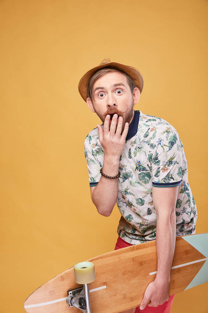 Surpised young man with handlebar mustache dressed in tropical print polo shirt, red shorts and straw hat, with longboard in hand. Studio shot with yellow background  - Foto, Imagen