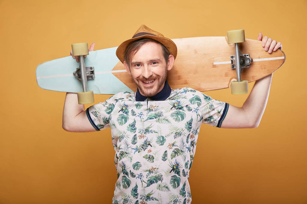 Cheerful young man with handlebar mustache dressed in tropical print polo shirt and straw hat, holding longboard behind head. Studio shot with yellow background  - Фото, изображение