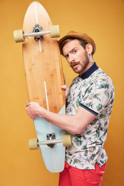 Young man with handlebar mustache and raised eyebrow, dressed in tropical print polo shirt, red shorts and straw hat, with longboard in hands. Studio shot with yellow background  - Foto, afbeelding