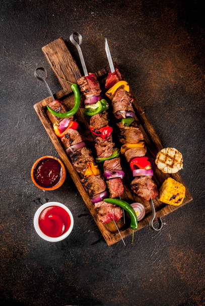 Fresh, home-cooked on the grill fire meat beef shish kebab with vegetables and spices, with barbecue sauce and ketchup, on a dark background on a wooden cutting board above copy space - Photo, Image