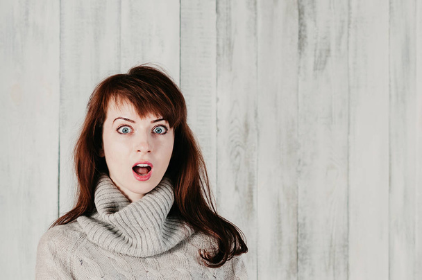 Een mooie jonge brunette vrouw in een grijze pullover, verrast grote ogen, open mond, met lichte achtergrond - Foto, afbeelding