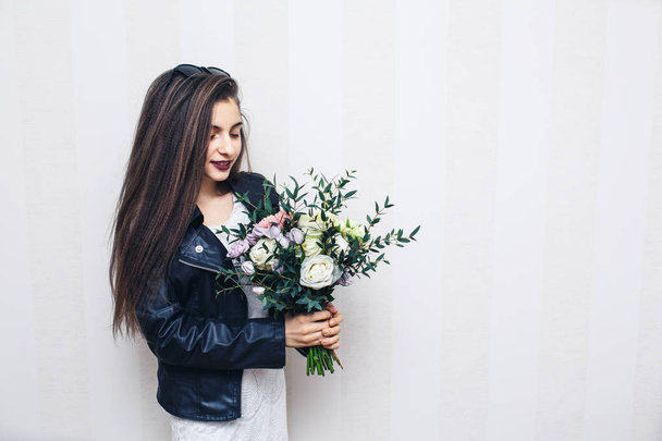 Beautiful stylish girl dressed in leather jacket, holding bouquet of flowers and posing against white wall - Foto, Bild
