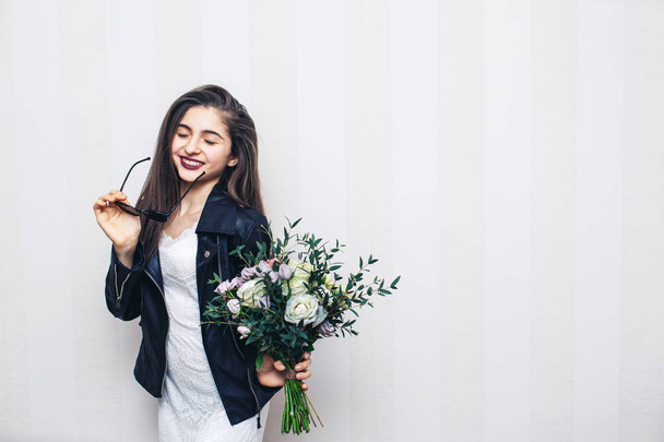 Beautiful stylish girl dressed in leather jacket, holding bouquet of flowers and posing against white wall - Photo, Image