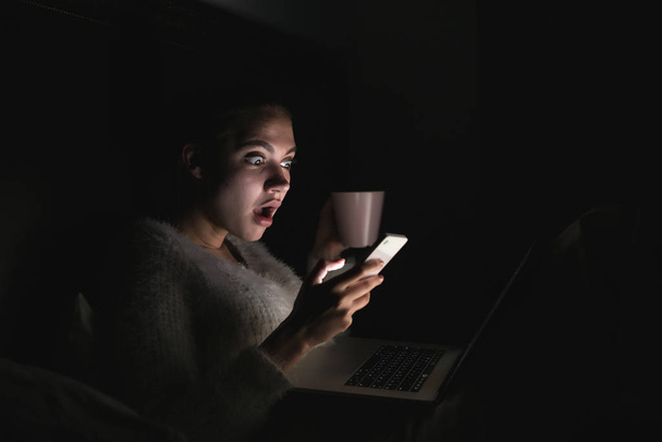 young surprised girl sitting in bed in the evening and watching a scary movie on her laptop - Fotografie, Obrázek