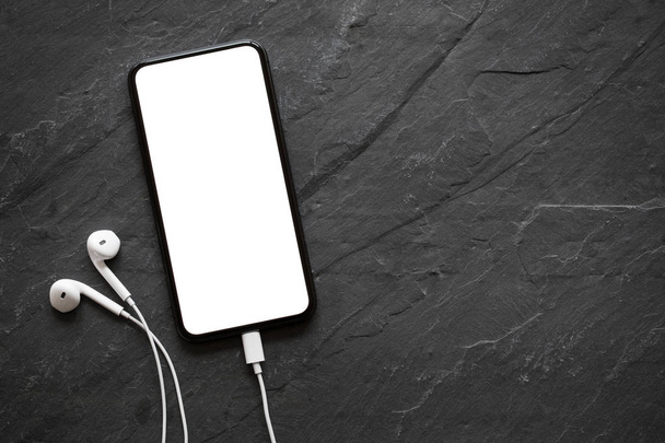 Мобільний телефон з порожнім екраном та навушниками на поверхні чорного каменю
 - Фото, зображення