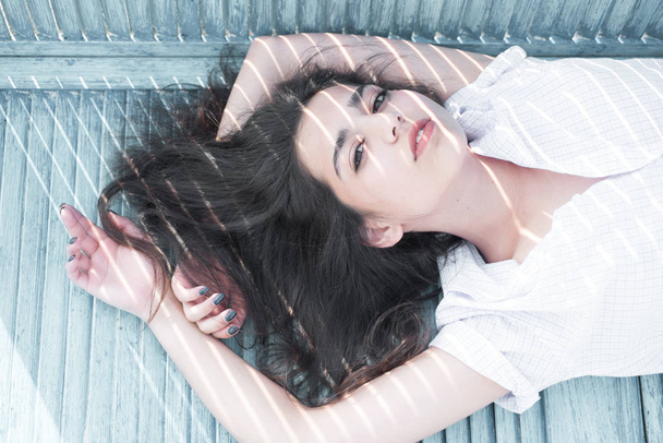 Woman in white outfit lying on floor, sensuality concept. Sexy brunette with long healthy hair on wooden balcony. Young pretty girl hiding in shade on hot summer day. - Foto, imagen