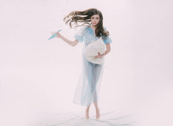 Charming girl with long wavy hair tiptoeing and throwing feathers in air isolated on ivory background. Slim young female holding big pillow and knife in hands, double nature, good vs evil concept. - Photo, image